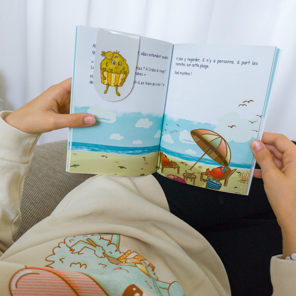 Enfant qui utilise le marque page cupcake momote sur le l'histoire de Crabe le rouge et les momotes à la plage.
